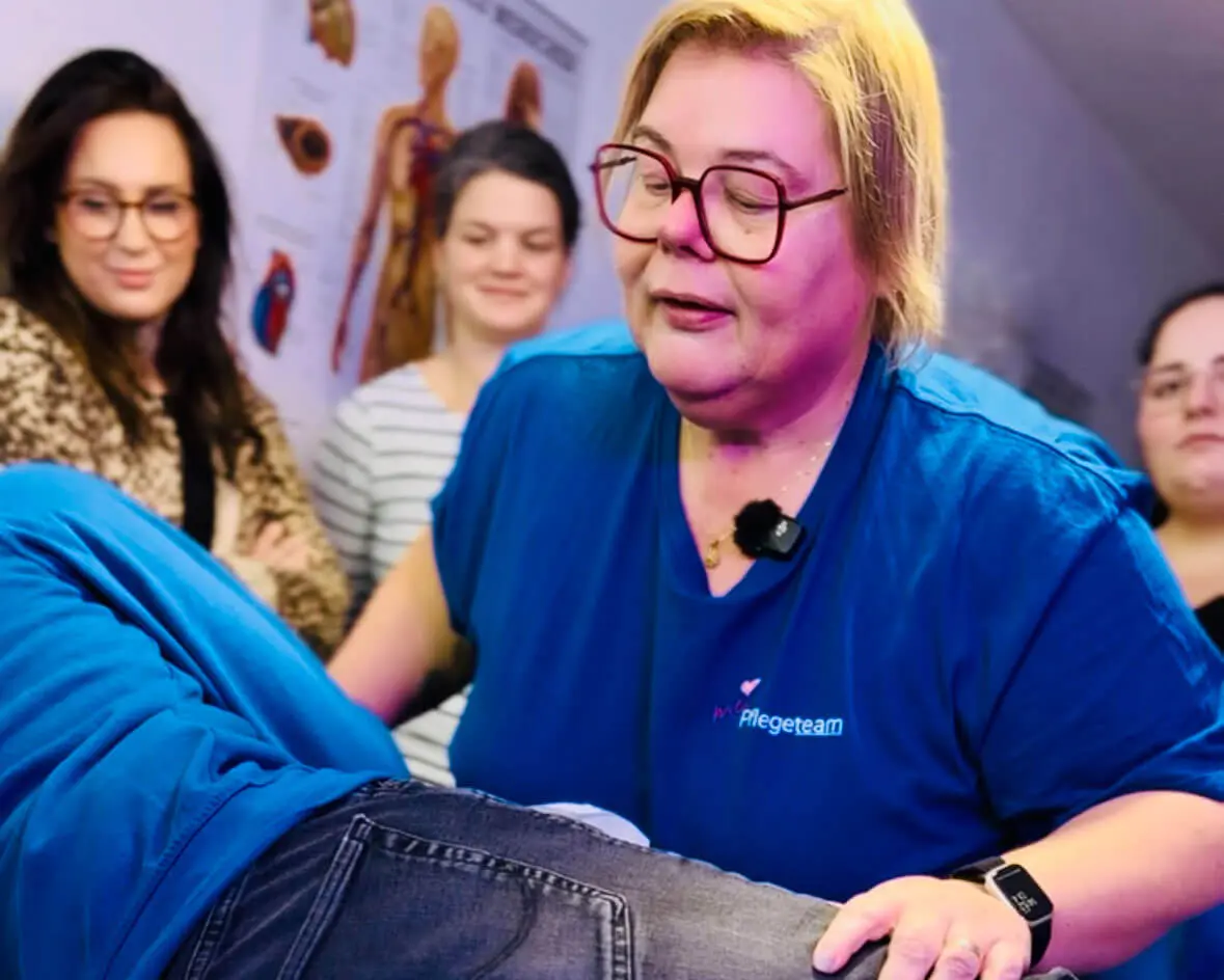 Pflegekraft in blauer Arbeitskleidung hält eine Person in Jeans liegend, im Hintergrund mehrere sitzende Personen vor anatomischen Postern