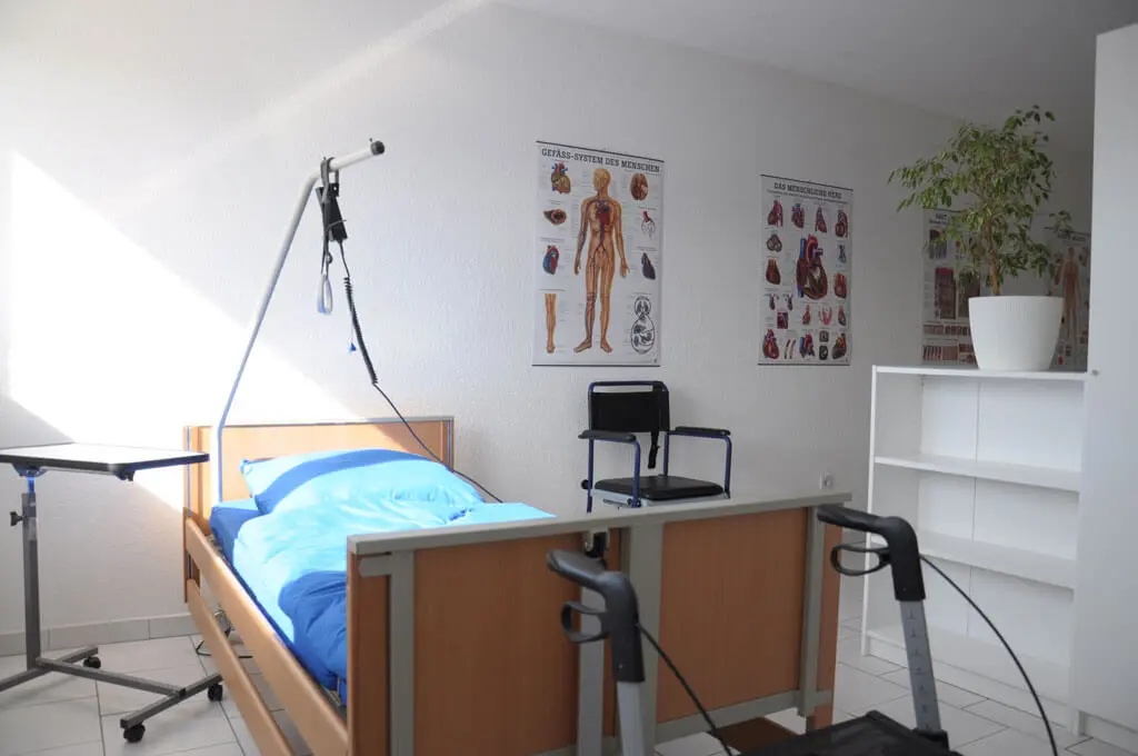 Schulungszimmer mit Pflegebett, Aufrichthilfe, Rollstuhl, Gehhilfen, anatomischen Lehrtafeln und einem Regal mit Pflanze