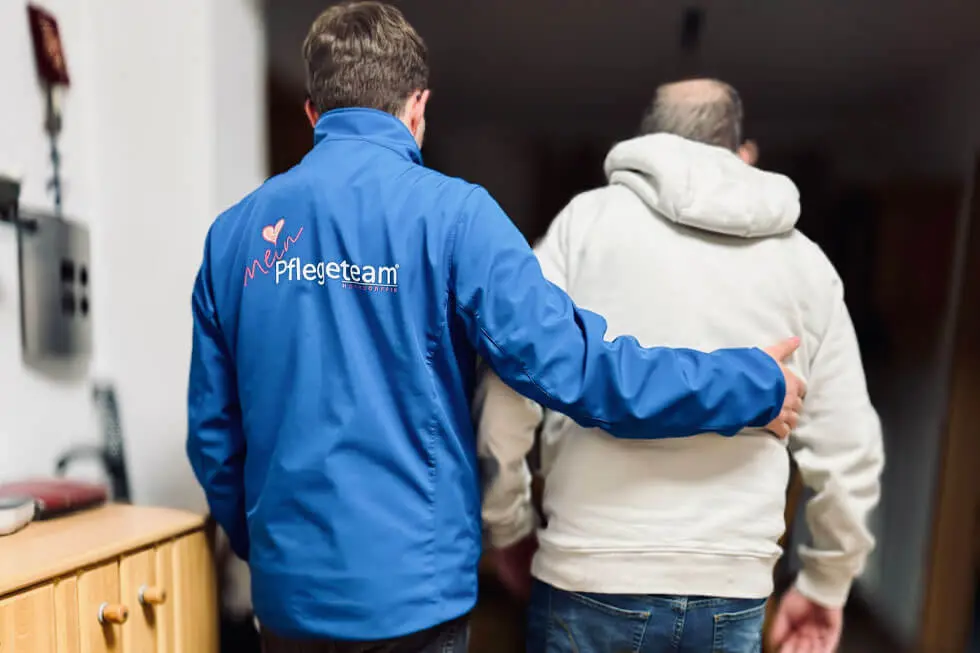 Pflegekraft in blauer Jacke mit Schriftzug 'Pflegeteam' stützt älteren Mann in weißer Kapuzenjacke von hinten beim Gehen