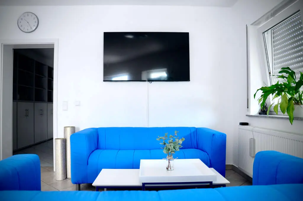 Wartebereich mit blauen Sofas, weißem Tisch, großer Pflanze am Fenster und an der Wand montiertem Flachbildschirm