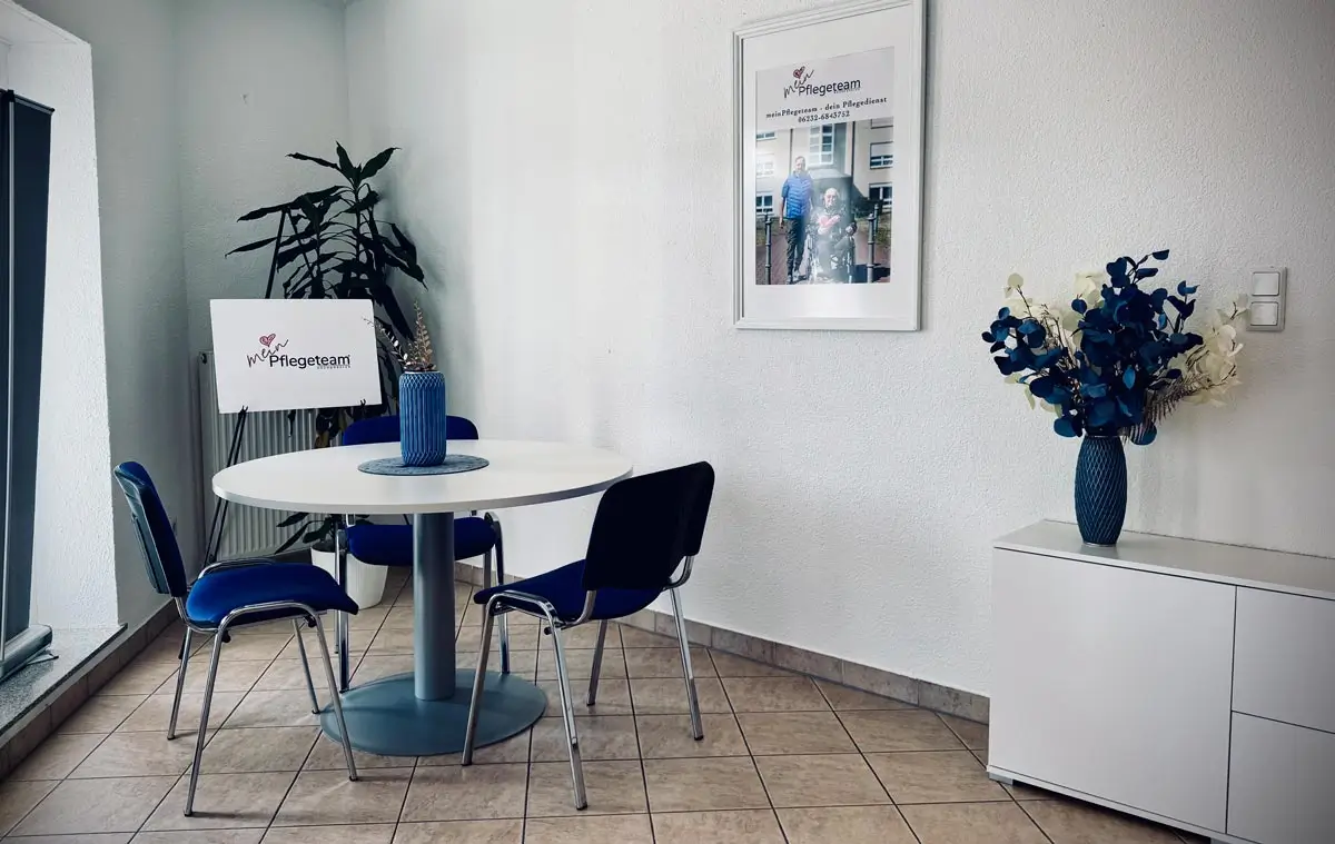 Beratungsbereich mit rundem Tisch, blauen Stühlen, Logo-Schild und Blumendekoration vor einer weißen Wand