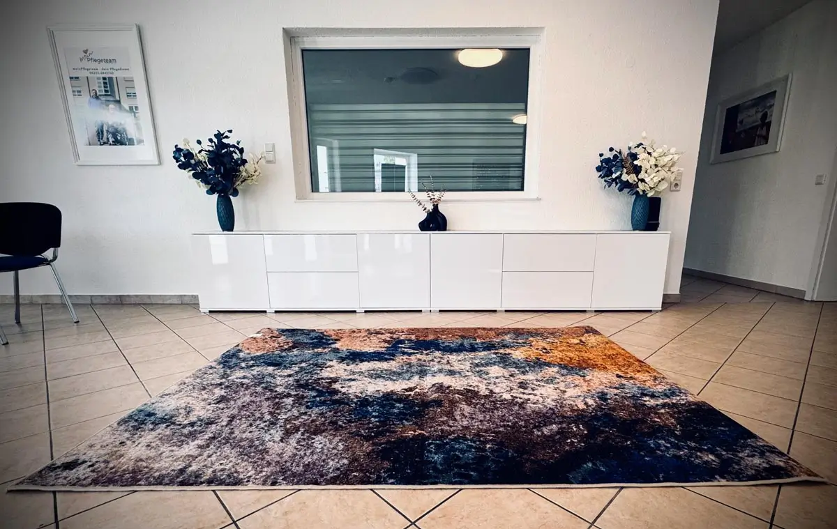 Empfangsbereich mit großem gemusterten Teppich, weißen Sideboard, Fenster zum Büro und Blumengestecken in blauen Vasen.