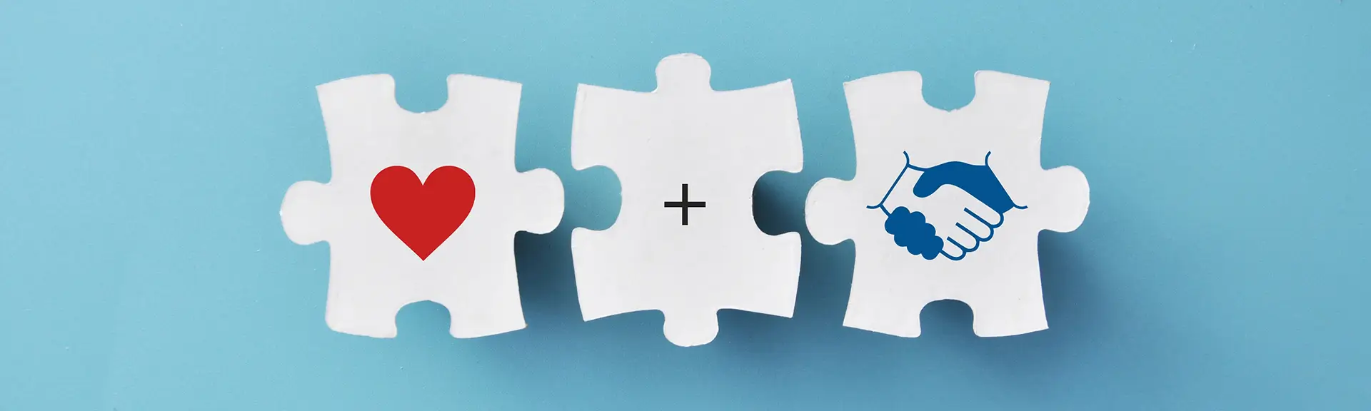 Drei weiße Puzzleteile auf blauem Hintergrund mit einem roten Herz, einem Pluszeichen und einem Handschlag