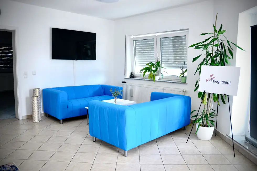Wartebereich mit zwei blauen Sofas, einem großen TV-Gerät an der Wand, Pflanzen auf der Fensterbank und einem Schild mit der Aufschrift 'Pflegeteam' neben einer großen Pflanze auf einem Ständer