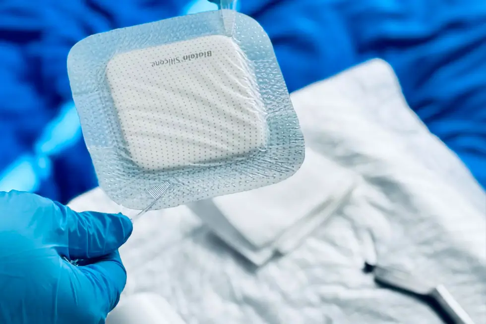 Hand mit blauem Handschuh hält steril verpacktes quadratisches Pflaster vor unscharfem medizinischem Hintergrund