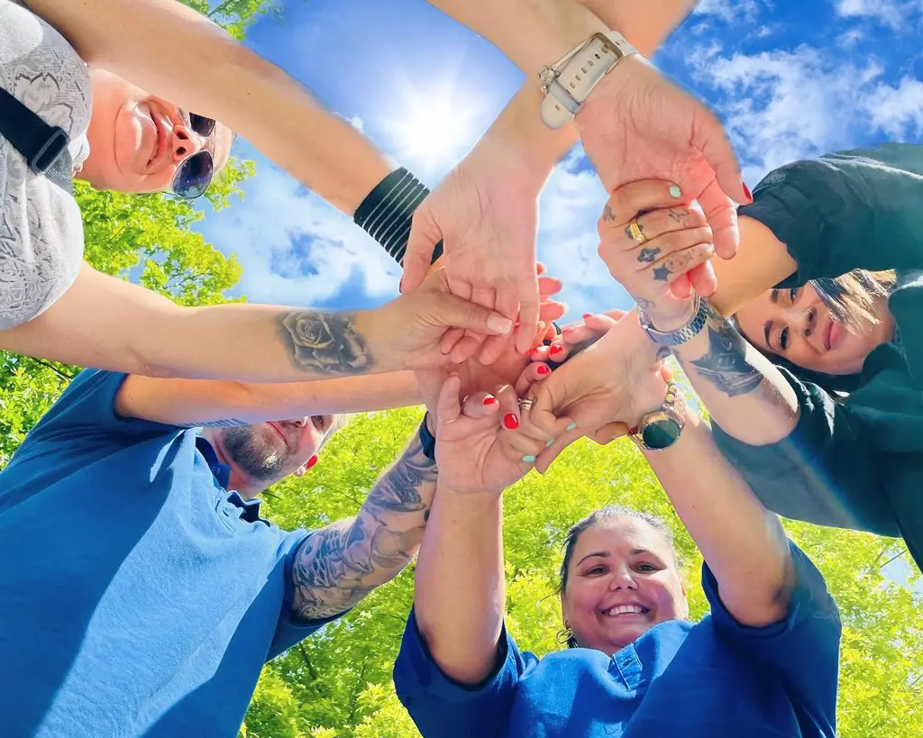 Mehrere Menschen stehen im Kreis und legen ihre Hände übereinander vor blauem Himmel und grünen Bäumen