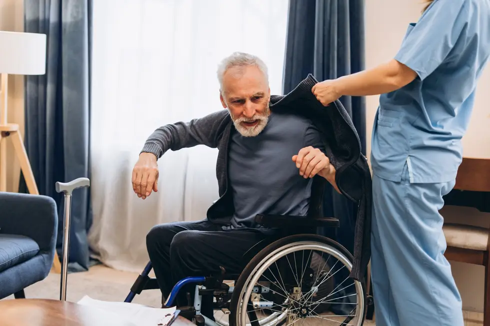 Pflegekraft hilft einem Mann im Rollstuhl beim Anziehen einer Jacke