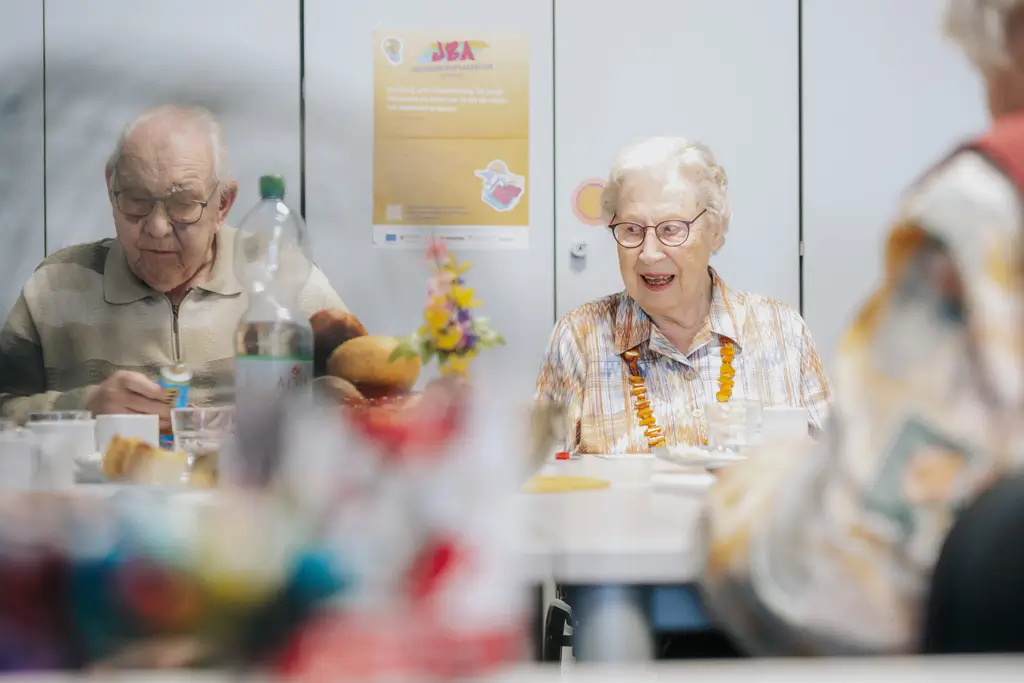 Zwei ältere Menschen sitzen an einem Tisch in einem Raum mit einem Poster an der Wand