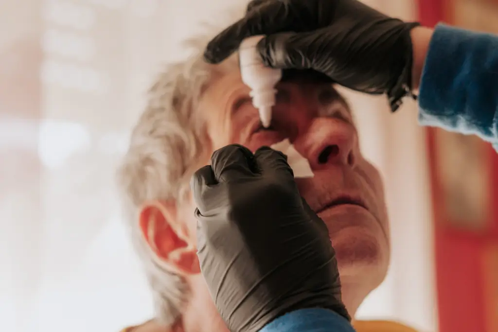 Person mit grauen Haaren erhält Augentropfen von einer anderen Person mit schwarzen Handschuhen