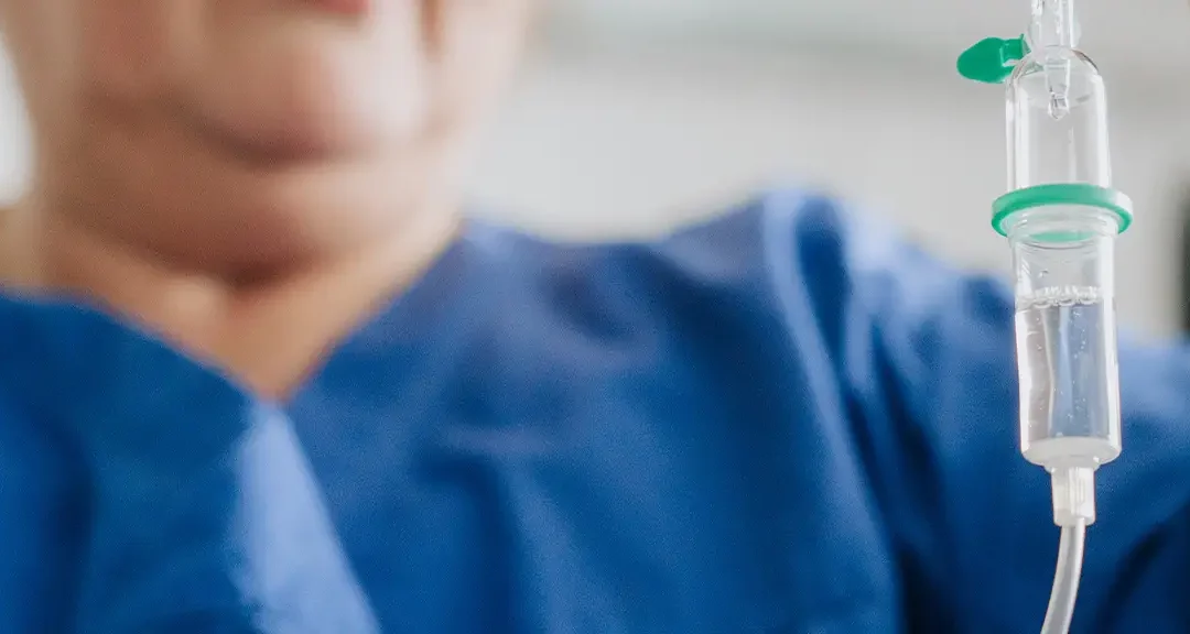 Person in blauer Arbeitskleidung hält Infusionsflasche mit Tropfkammer in der Hand