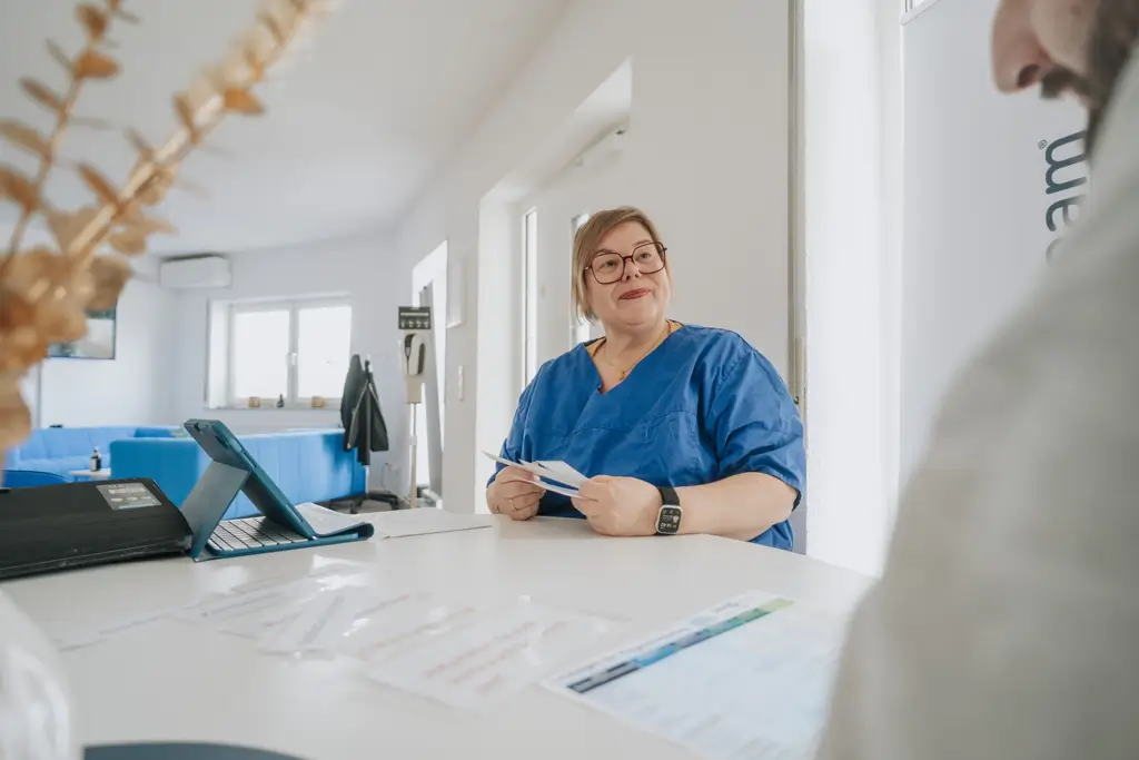 Person in blauer Arbeitskleidung sitzt an einem Tisch und hält Papiere in der Hand, im Vordergrund unscharf eine weitere Person mit Dokumenten