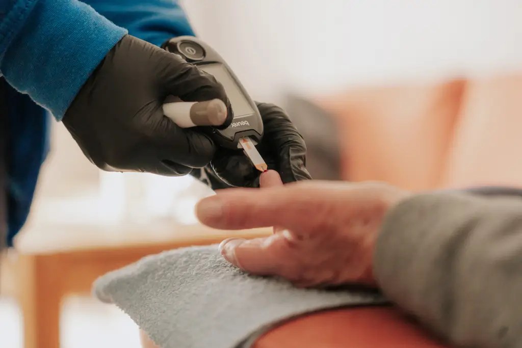 Hand mit schwarzem Handschuh hält Blutzuckermessgerät an Finger einer anderen Person