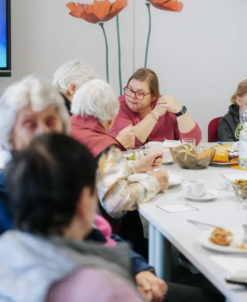 Mehrere Personen unterschiedlichen Alters sitzen an einem gedeckten Tisch und unterhalten sich, im Hintergrund sind große Wandbilder von Mohnblumen zu sehen.