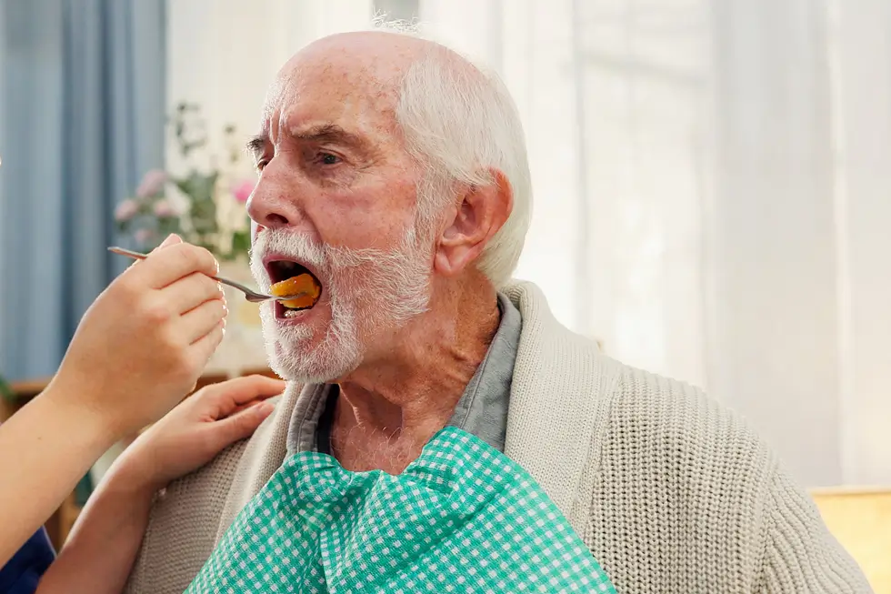 Eine Frau füttert einen älteren Mann, der eine grün-weiß karierte Lätzchen trägt, mit einem Löffel
