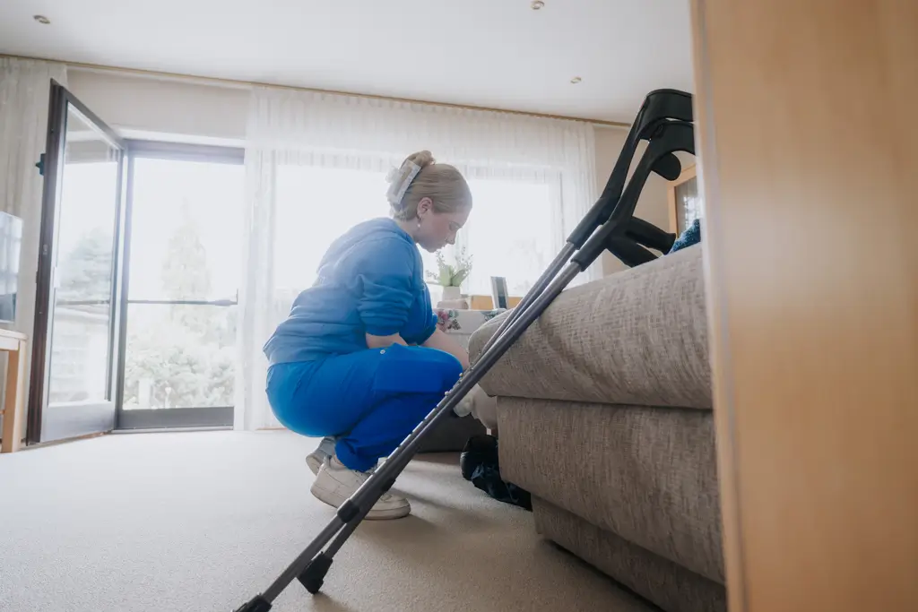 Person in blauer Arbeitskleidung kniet vor einem Sofa und reinigt den Boden, im Vordergrund steht ein Staubsauger