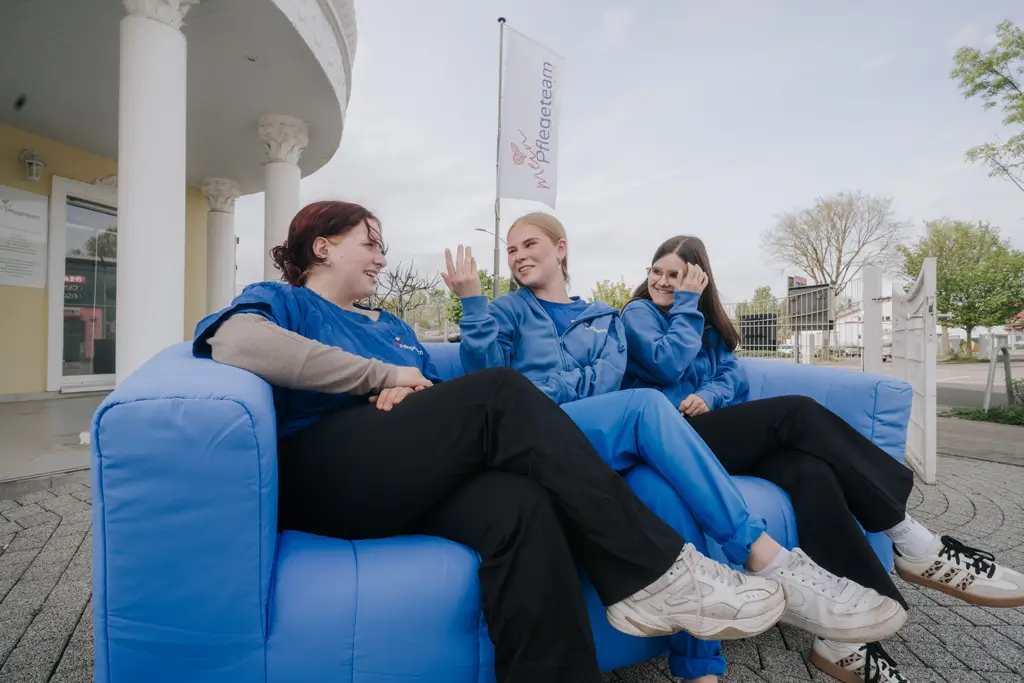 Drei Personen in blauen Arbeitskleidungen sitzen auf einem blauen Sofa im Freien vor einem Gebäude mit einer Fahne im Hintergrund