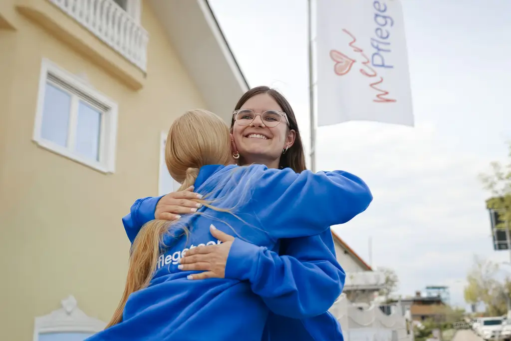 Zwei Personen in blauen Pullovern umarmen sich vor einem Gebäude mit einer Fahne, auf der 'mari Pflege' steht
