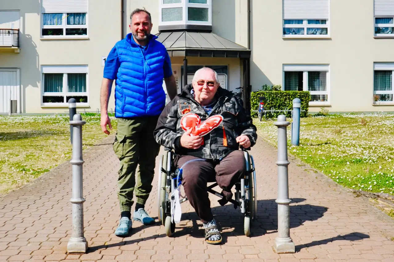 Ein Mann schiebt einen Rollstuhlfahrer auf einem gepflasterten Weg vor einem Wohngebäude