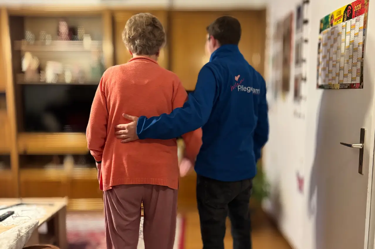 Pflegekraft mit blauem Pullover stützt ältere Frau mit oranger Jacke beim Gehen in einer Wohnung