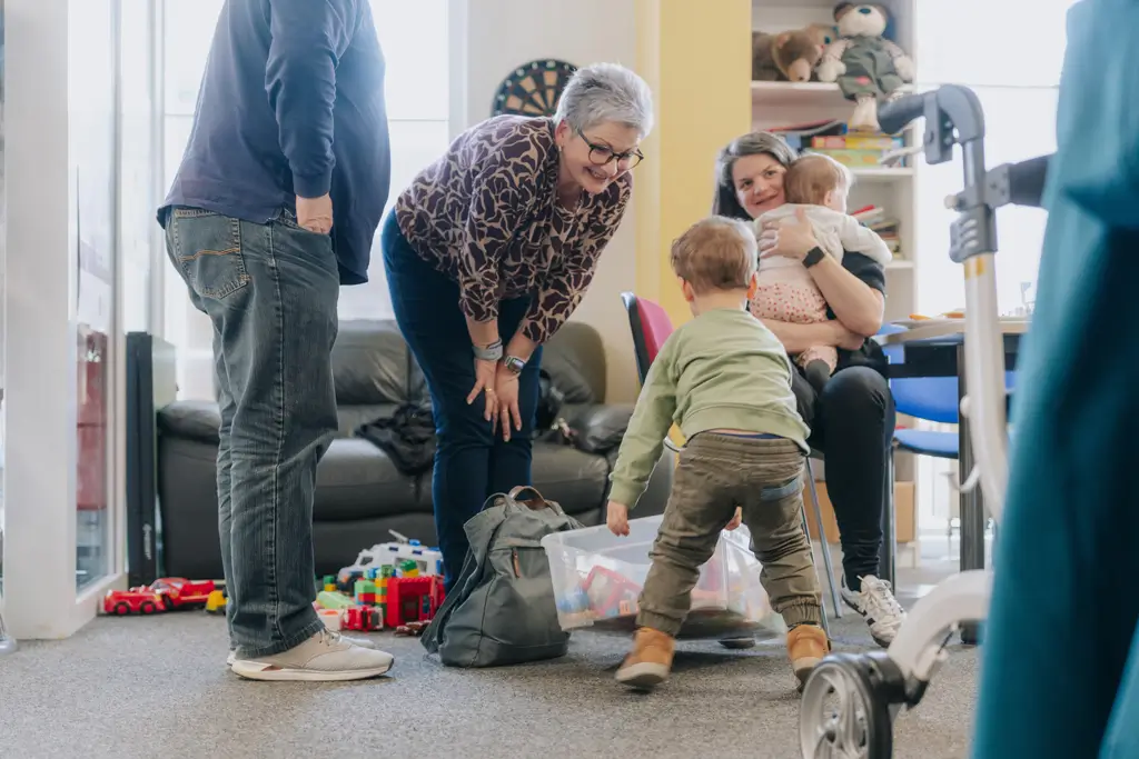 Mehrere Personen in einem Wohnzimmer, darunter ein Kind, das sich zu einer Kiste mit Spielzeug hinunterbeugt, eine ältere Frau, die sich vorbeugt, und eine sitzende Frau, die ein Baby hält
