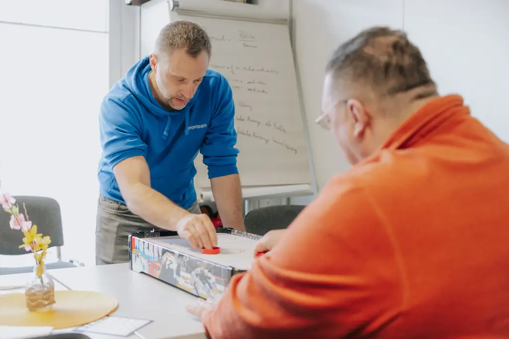 Zwei Männer spielen Tischhockey an einem Tisch, einer trägt einen blauen Kapuzenpullover, der andere ein oranges Oberteil