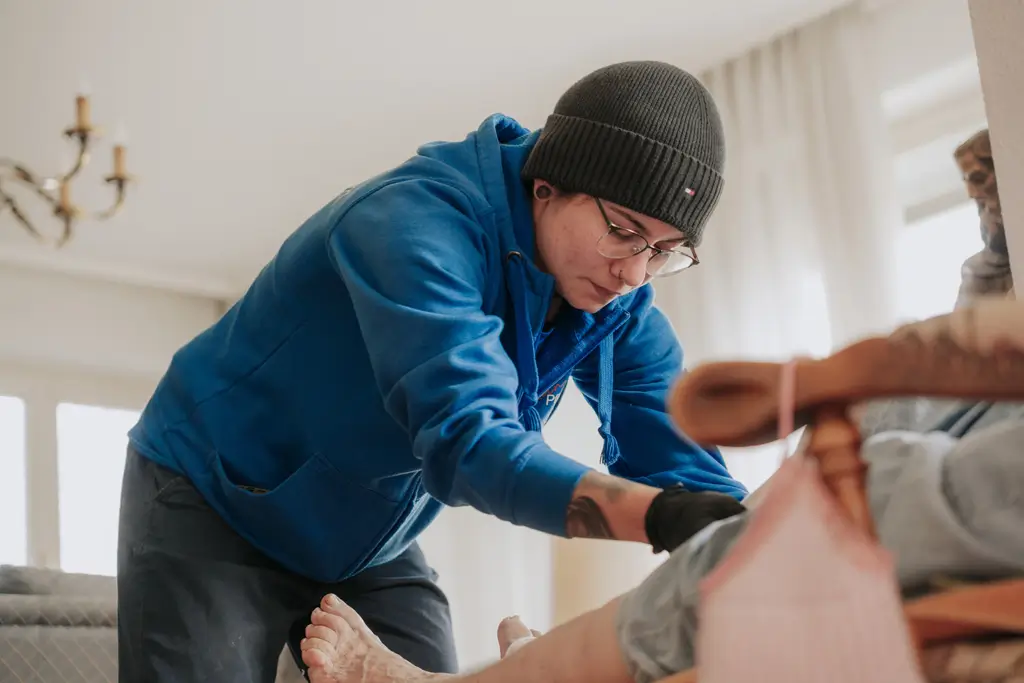 Person in blauer Kapuzenjacke und Mütze beugt sich über eine sitzende Person und berührt deren Bein