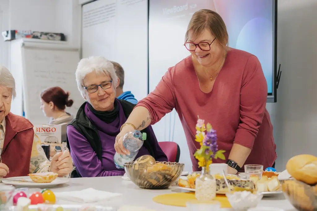 Frau gießt Wasser aus einer Flasche in eine Schüssel auf einem gedeckten Tisch mit mehreren Personen im Hintergrund