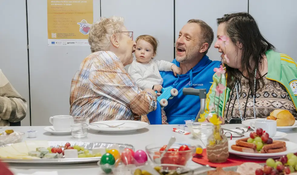 Vier Personen unterschiedlichen Alters sitzen an einem Tisch und unterhalten sich, darunter ein älterer Mann, ein Kleinkind, ein Mann in blauer Kleidung und eine Frau mit langem dunklem Haar
