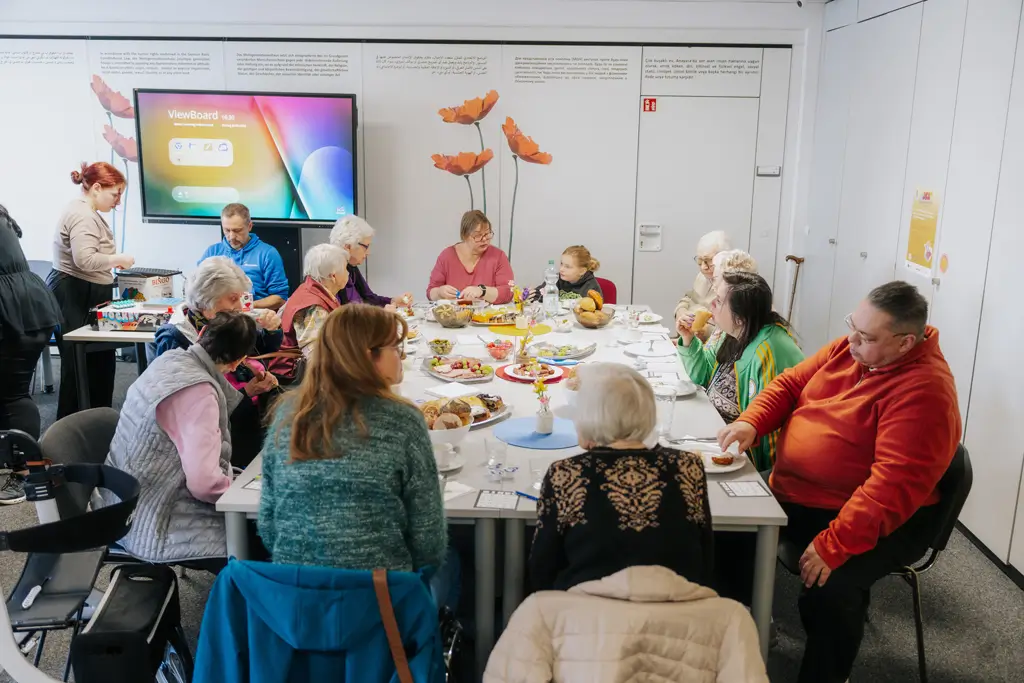 Gruppe von Menschen unterschiedlichen Alters sitzt an einem langen Tisch und isst gemeinsam in einem hellen Raum mit weißen Wänden und einem Bildschirm an der Wand