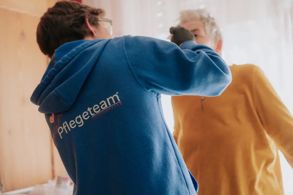Pflegekraft in blauem Pullover mit Aufschrift "Pflegeteam" versorgt ältere Person in gelbem Pullover