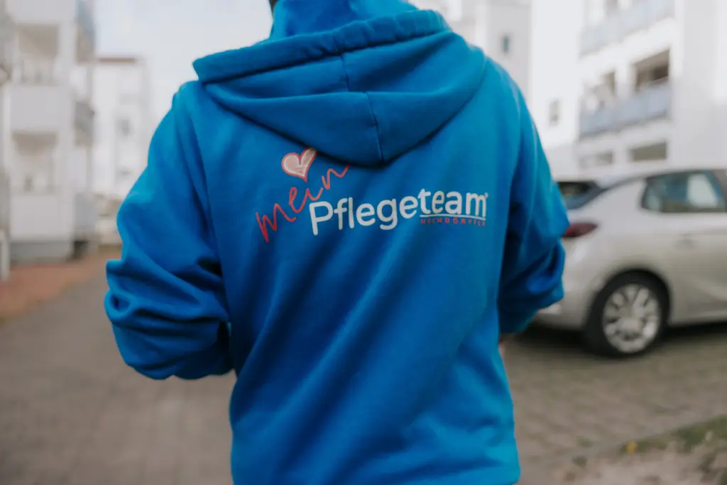 Person von hinten mit blauem Kapuzenpullover und Aufschrift 'mein Pflegeteam' auf dem Rücken in einer Wohnsiedlung
